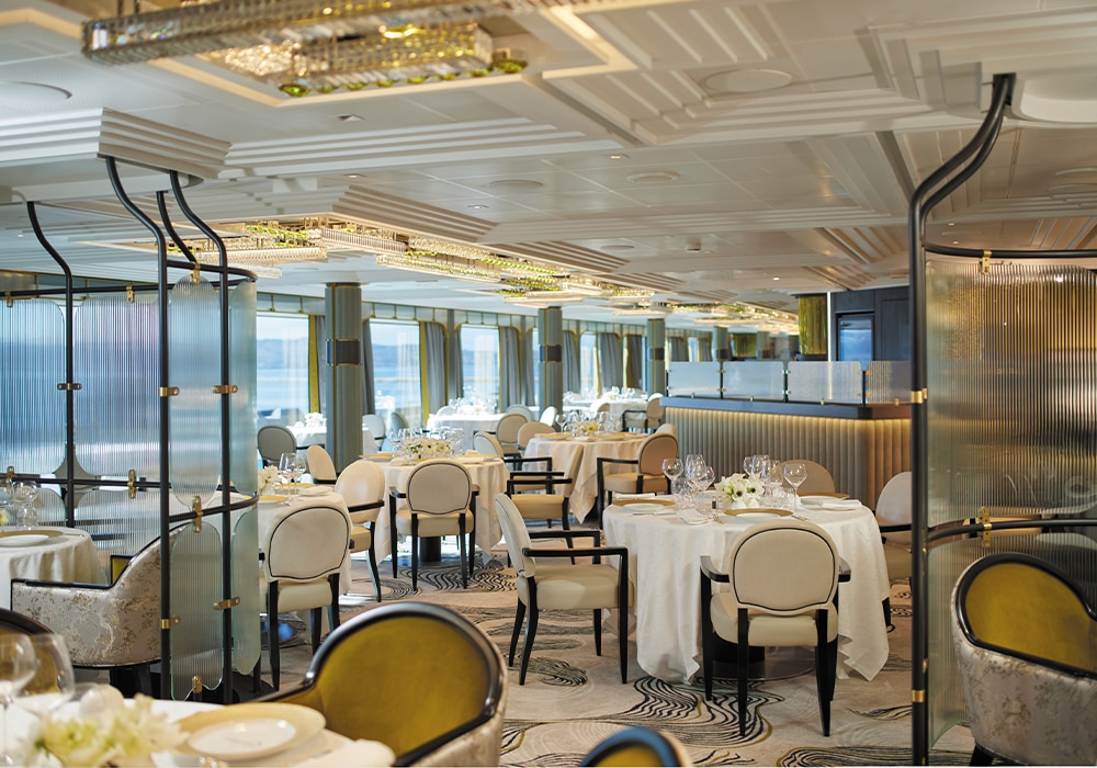Lichte eetzaal aan boord met ronde tafels, crème fauteuils en grote ramen op zee